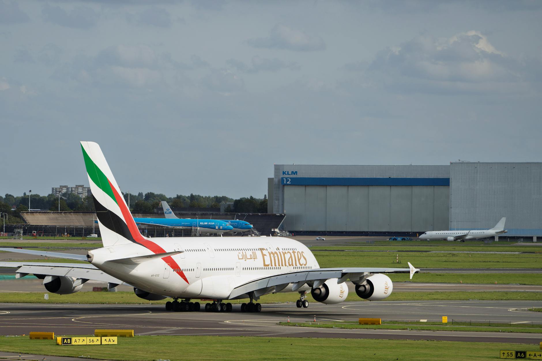 An Emirates A380 aircraft taxiing on a runway with KLM hangar in the background.