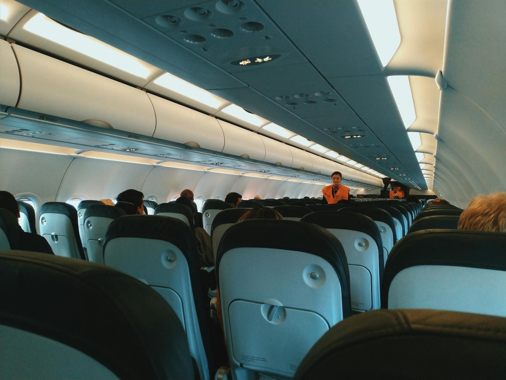 Inside of modern airplane cabin with passengers sitting on comfortable seats and cabin crew standing at passageway