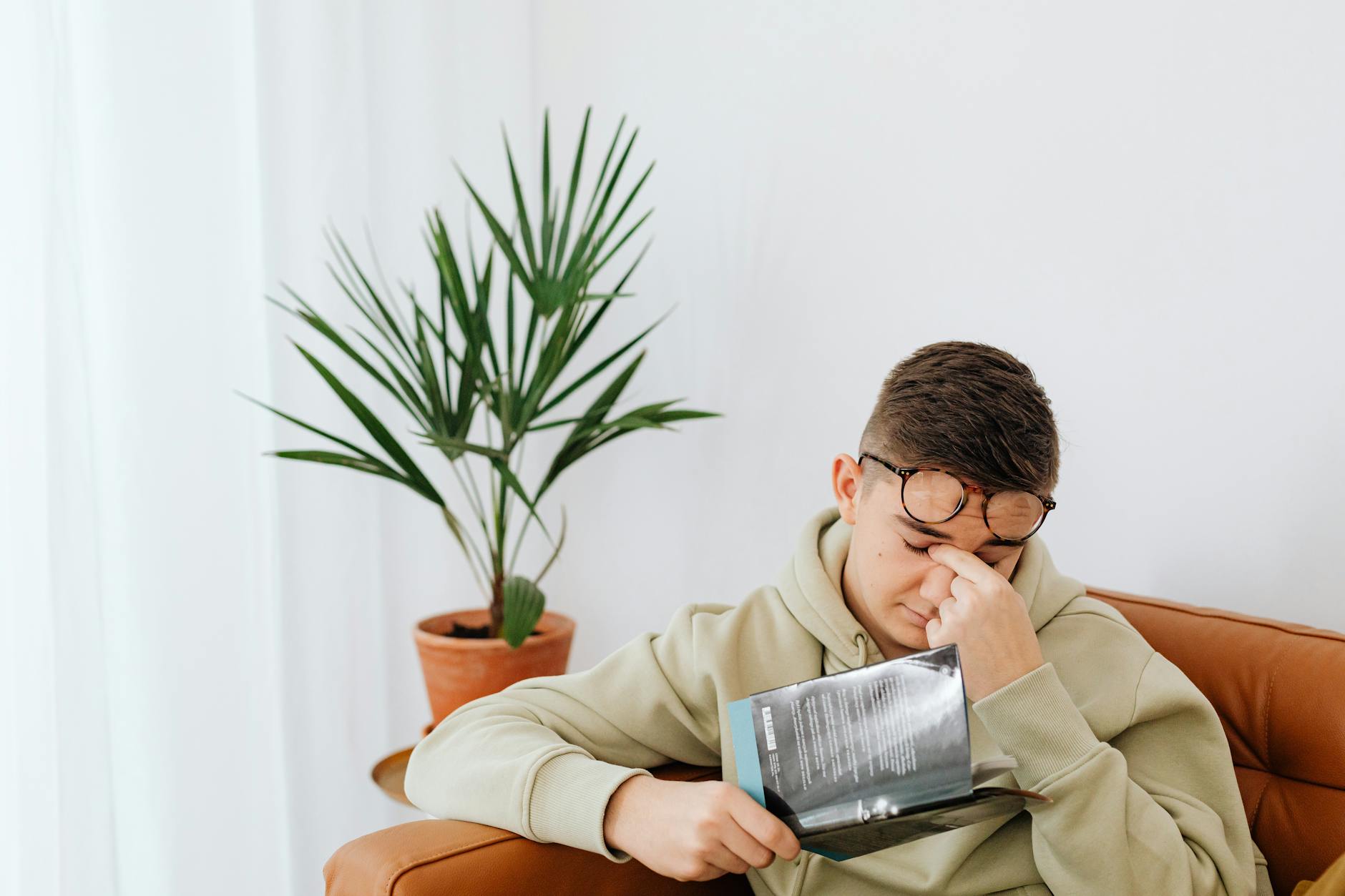 Adult man wearing hoodie rubs his eyes while reading a book indoors, next to a potted plant.